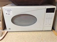 Front view of white GE tabletop microwave showing oval window door, digital clock on keypad panel, and maxsold tag #380