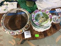 Photo showing large glass serving bowl with ceramic floral rim next to a stack of assorted decorative plates with tropical and floral patterns on a wooden table