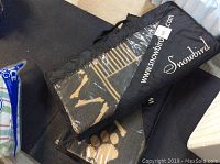 Two black Snowbird brand outdoor rugs in carry cases placed on a floor rug, cases have clear plastic windows showing beige patterned rugs inside. Cases have white website URL and brand name.