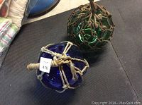 Two antique Japanese glass fishing net floats, blue and green, with beige rope netting on a black mat.