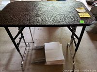 Photo of adjustable folding table with black textured top, foldable metal legs, and miscellaneous papers underneath.
