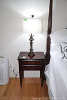 Full shot showing the Bombay side table next to a bed with a lamp on top, demonstrating size and style.