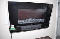 Front view of black wall mounted electric fireplace with glass front and faux log detail inside. Power cord visible.
