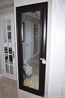 Tall wood framed mirror hanging on a wall reflecting door and hallway.
