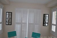 Two matching decorative wall mirrors hanging on either side of a window with white shutters, each mirror having a black frame and geometric grid pattern overlay on the glass.