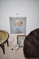 Five framed beach-themed art pieces arranged leaning against a wall near a brown chair and round wooden table. Large ammonite shell and coral print visible.