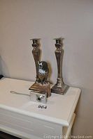 Two tall silver candle holders with classical design, owl figurine on wood base, and silver Sony Cybershot camera on white surface against wall.