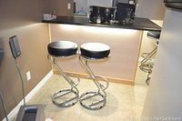 Two black and chrome bar stools positioned by a countertop in a room with beige tile flooring and brown walls.
