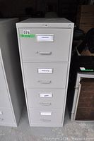 Front view of the grey metal filing cabinet with all four drawer fronts visible, showing label holders and recessed handles.