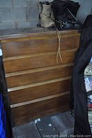 Wooden dresser with five drawers shown from front, medium-tone wood, placed against a wall in a storage area.