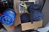 Three sleeping bags in a cardboard box and on top: two dark blue sleeping bags, one rolled and strapped with a brown belt, the other folded, and one blue rolled sleeping bag with black straps