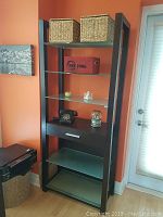 Full view of dark wood frame shelving unit against orange wall, showing six glass shelves and top baskets