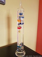 Full view of the 14-inch Galileo thermometer with colorful floating spheres inside a clear glass cylinder on a glass base.