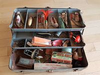 Full view of opened gray metal tackle box with fishing lures, reel, lure boxes, and fishing accessories organized in compartments.