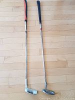 Two mallet head putters shown side by side on wooden floor, one with black grip and one with orange and black grip
