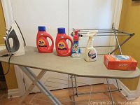 Sunbeam steam iron, detergent bottles, ironing board, drying rack, and laundry supplies displayed on ironing board and floor.