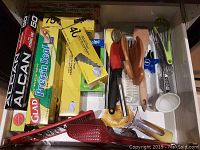 Photo of kitchen drawer showing boxes of plastic wrap, aluminum foil, parchment paper, and freezer bags alongside wooden handled utensils and kitchen tools.