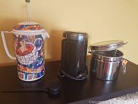 All three items displayed side by side: mosaic coffee press, black electric grinder, and stainless steel canister with clamp lid and scoop on top.