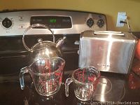 Photo showing stovetop with stainless steel kettle, two glass measuring cups, and Cuisinart stainless steel two-slice toaster