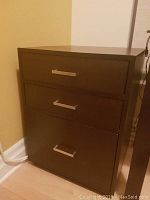 Full view of dark brown solid wood three drawer filing cabinet with silver handles, placed in corner against wall and floor baseboard.