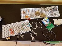 Full overview of multiple pairs of earrings and pins arranged on black surface with white mat, showing variety of designs including dangling beads, hoops, and pins.