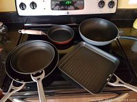 Image showing three pans on stove including wok with lid, small frying pan, and square grill pan with ribbed surface.