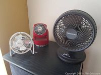 Three fans on black table: white Breeze'n In, red Vornado, black Sunbeam Blizzard