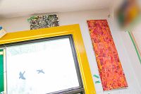 Photo showing two of the paintings hung above a window with yellow trim; one painting predominantly red and pink vertical rectangle and partially visible abstract painting with green and white swirling pattern