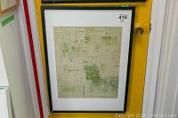 Framed abstract artwork hanging against a yellow door, showing the full piece with a black frame and white matting.