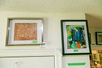 Two framed original abstract paintings hanging on a wall, one with earthy warm tones in a light frame, the other colorful with a dark frame.