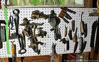 Pegboard with plumbing fixtures, valves, faucets, and used pocket knives hung alongside small hand tools including a hand drill and brushes