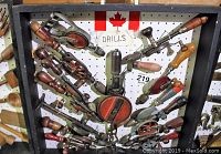 View of up to 15 manual crank hand drills mounted on a black open pegboard case with a Canadian flag and 'DRILLS' sign above them
