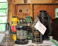 Seven lanterns displayed on wooden shelf, showing variety of colors and designs including ribbed glass and wire cage lanterns, and a small pressed metal patterned lantern shade in front.