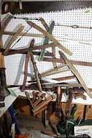 Overview of vintage cross-cut saws and buck saws with wooden clamps arranged in front of pegboard.