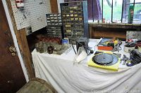 Wide view showing the table covered with a white cloth holding multiple plastic drawers with screws, jars with screws, hand tools, saw blades, and various small parts.