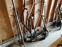 Garden tools leaning against a wooden wall showing multiple shovels, a broom, and scythe
