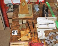 Photo showing wooden metronome, ink well, lighters, two Sheaffer boxed pens, key chains, coins and Bolivian banknote on a wooden table.