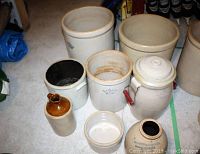 Set of eight stoneware crocks and containers arranged on floor showing various sizes, some with lids and blue crown emblems visible