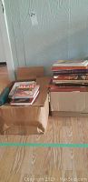 Two cardboard boxes filled with books placed on a wooden floor, showing the volume and types of books included.