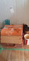 Folded orange and white picnic blankets with palm tree design stacked on floor with some travel mugs beside them