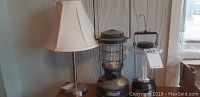 Lot containing two electric lanterns and one table lamp with fabric shade, shown against a wall and sitting on wooden surface