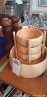 Four medium wooden salad bowls nested vertically in a wooden stand with one larger wooden salad bowl around the base, salt and pepper shaker grinder behind