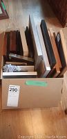 Box of assorted empty frames in various sizes and styles stacked vertically in a cardboard box on a wooden floor.