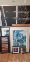 Framed prints and photographs arranged leaning against a wall including a large black and white bridge photo, blue village scene print, multicolored abstract print, and smaller framed pieces
