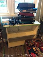 Wide shot of white three-tiered shelf with stacked binders and electronics on top, with visible dust and dirt on shelf surfaces.