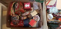 Photo of a box with assorted Christmas tins, decorative ribbons, and small gift boxes with holiday patterns.