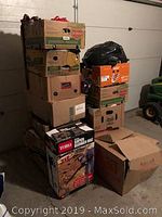 Stack of 13 cardboard boxes and one black plastic bag containing assorted clothing and shoes, shown in garage