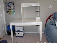 Front view of white Ikea desk with removable shelf, placed in a bedroom next to a blue chair and wall decorations.