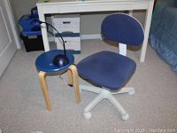 Blue rolling office chair, blue round stool table with wooden legs and blue dome-shaped lamp on top, all placed on carpeted floor near white desk