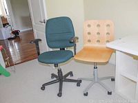 Two rolling office chairs side by side, one teal padded fabric chair with armrests and black base, and one light wood chair with circular backrest cutouts on grey base.